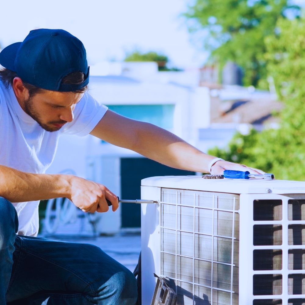 Technicien portant une casquette bleue et utilisant un tournevis pour réparer un climatiseur sur le toit.